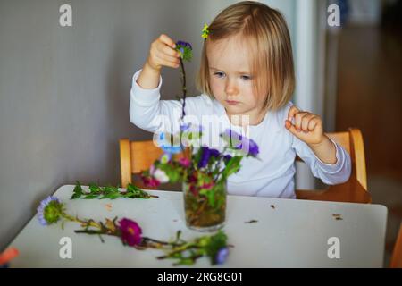 Adorabile bimba che prepara il bouquet di fiori a casa. Bambino che mette i pani in acqua. Attività domestiche per bambini Foto Stock