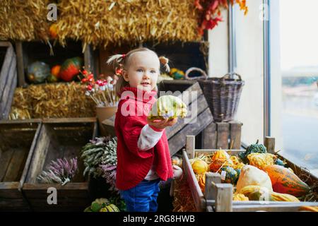 Adorabile bambina con poncho rosso che seleziona zucche colorate nel supermercato, nel negozio di alimentari o in fattoria. Felice ragazzo che festeggia Halloween Foto Stock