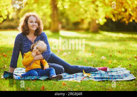 Donna e adorabile bimba seduta a terra per fare un picnic nel parco autunnale. Madre e figlia si godono il sole della giornata d'autunno insieme. Esterno Foto Stock