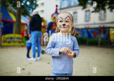 Piccola bambina in età prescolare con un gatto che dipinge il viso, che fa una buffa schifosa all'aperto. I bambini dipingono il viso. Attività creative per bambini Foto Stock