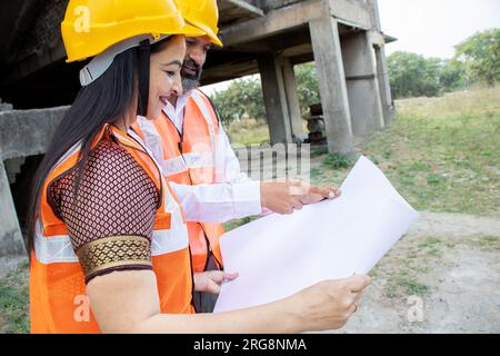 Due ingegneri civili indiani, uomini e donne, o architetti, che indossano casco e giubbotto con un progetto di scartoffie in cantiere che discutono di una vera e propria proprietà Foto Stock