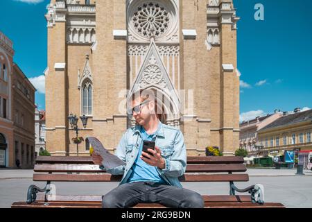 Uomo turista siede su una panchina nella piazza della città con mappa e cellulare e cerca l'hotel che ha prenotato tramite un'applicazione sul suo telefono viaggiatore maschio Foto Stock