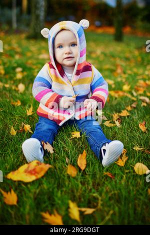 Adorabile bambina seduta sull'erba e giocando con le colorate foglie autunnali in una giornata autunnale nel parco Foto Stock