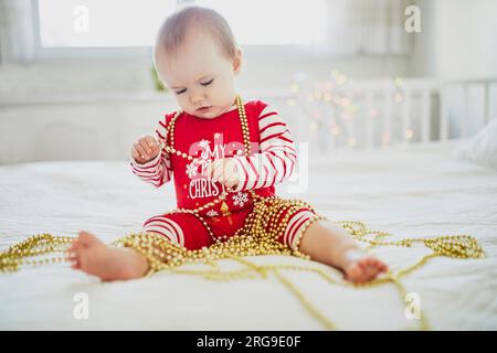 Bambina felice che indossa un pigiama che gioca con le decorazioni dell'albero di Capodanno nel suo primo Natale. Festeggiamo il Natale con i bambini a casa Foto Stock