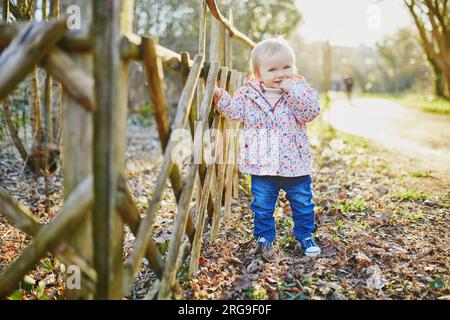 Una bambina di un anno in piedi accanto a una recinzione di legno nel parco. I bambini imparano a camminare. Foto Stock