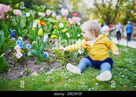 Una bambina di un anno seduta sull'erba con tulipani colorati. Bambino che guarda i fiori in una giornata primaverile nel parco. Adorabile bambino che esplora la natura Foto Stock