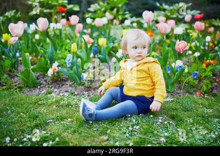 Una bambina di un anno seduta sull'erba con tulipani colorati. Bambino che guarda i fiori in una giornata primaverile nel parco. Adorabile bambino che esplora la natura Foto Stock