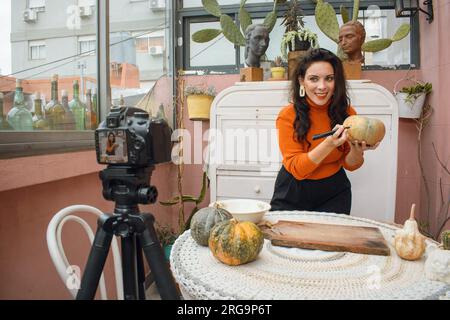 Creatrice di contenuti di donna caucasica che scatta una foto per i social network con la sua fotocamera digitale su un treppiede mentre sorride e disegna una zucca per Halloween Foto Stock