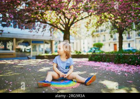 Adorabile bambina che disegna l'arcobaleno con gessetti colorati sull'asfalto nel parco con alberi di ciliegio in fiore. Attività all'aperto e giochi creativi Foto Stock