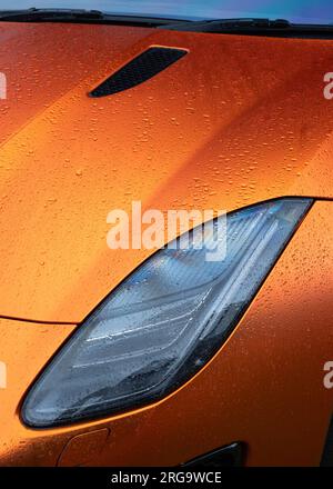 Un primo piano della sezione del faro anteriore sinistro di una vettura sportiva britannica Jaguar F-Type di colore arancione brillante coperta da gocce di pioggia Foto Stock