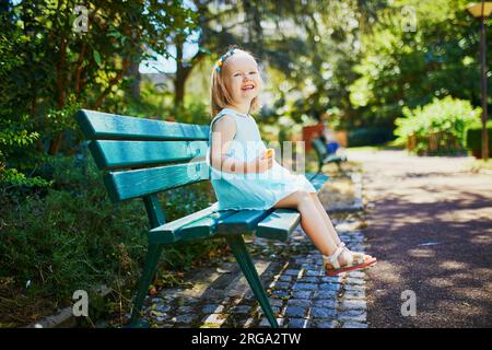 Adorabile bimba seduta sulla panchina all'aperto e che fa uno spuntino in una soleggiata giornata estiva. Un bambino felice che cammina nel parco. Passeggiate e alimentazione sana per Foto Stock