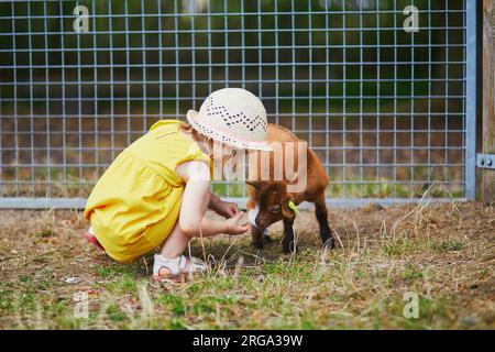 Adorabile bambina con abito giallo e cappello di paglia che gioca con le capre alla fattoria. Bambino che si familiarizza con gli animali. Agricoltura e giardinaggio per il SM Foto Stock