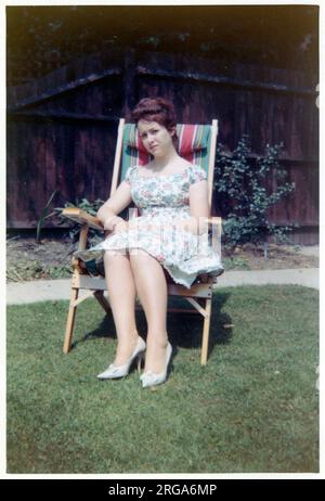 Una giovane donna che indossa un abito estivo con stampa floreale, sedette in un bel giardino in una sedia a righe con retro in tela. Foto Stock