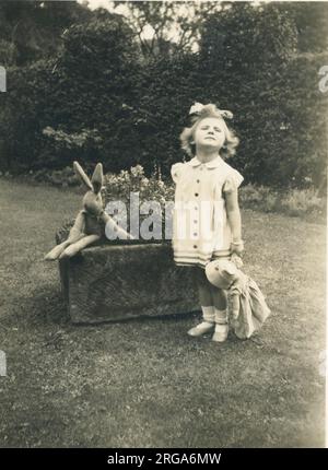 Una bambina si mette all'attenzione e si lamenta per la macchina fotografica in un giardino con in mano una bambola, mentre il suo coniglietto giocattolo si siede vicino. Foto Stock