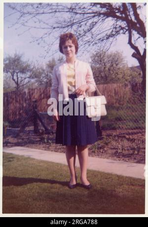 Una giovane donna che si posa per una fotografia in un giardino suburbano pulito. Indossa una blusa canarino giallo, un cardigan bianco (e una borsa abbinata) e una gonna scura intelligente - Agosto, 1960 Foto Stock