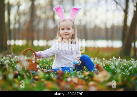 Ragazza che indossa orecchie da coniglio che gioca a caccia all'uovo a Pasqua. Bambino seduto sull'erba con molti fiori a goccia di neve e raccogliendo uova colorate nel cestino. Acceso Foto Stock