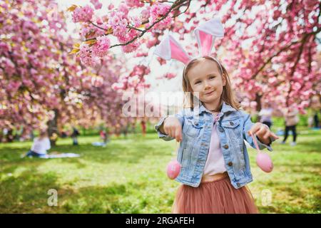 Bambina in età prescolare che indossa le orecchie del coniglio e gioca a caccia all'uovo a Pasqua. I bambini raccolgono uova colorate nel cestino. Bambino che festeggia la Pasqua all'aperto Foto Stock