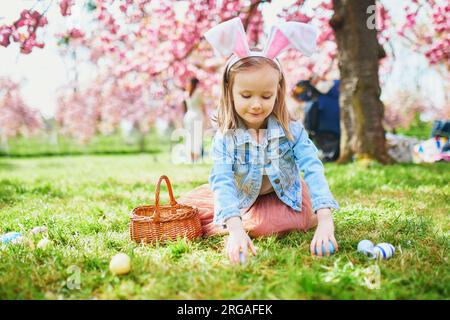 Bambina in età prescolare che indossa le orecchie del coniglio e gioca a caccia all'uovo a Pasqua. I bambini raccolgono uova colorate nel cestino. Bambino che festeggia la Pasqua all'aperto Foto Stock