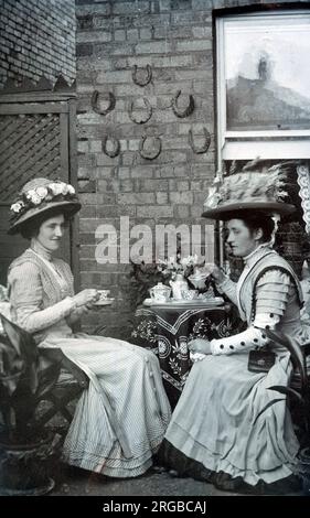 Due donne edoardiane di classe media prendono il tè in un giardino di periferia e concedono un po' di pettegolezzi leggeri. Foto Stock