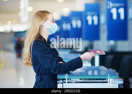 Giovane donna che indossa una maschera protettiva all'aeroporto internazionale al banco del check-in, dà il passaporto a un ufficiale e aspetta il suo imbarco Foto Stock