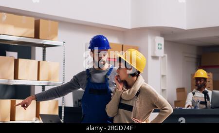 Team di persone che parlano dell'inventario in magazzino, della merce nei pacchi sugli scaffali dei magazzini. Diversi lavoratori in tute e hardHat che utilizzano un computer portatile per pianificare la spedizione degli ordini. Foto Stock