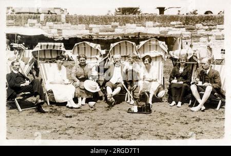 Un gruppo di famiglie si goda un po' di sole estivo e una tazza di tè sulla spiaggia (possibile nel Dorset? Costa meridionale dell'Inghilterra?). Hanno noleggiato eleganti sdraio, ognuna con ombrelloni pieghevoli! Foto Stock