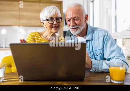 Allegra coppia anziana seduta al tavolo da pranzo con cucina in background e navigare su internet sul computer portatile Foto Stock