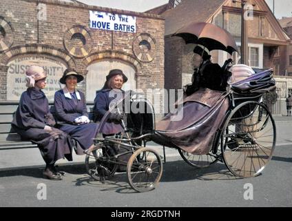 Scena di strada vicino alle Terme di Hove in Sussex. Tre donne si siedono su una panchina, vicino ad una donna anziana in un'elaborata sedia da bagno a quattro ruote, ombreggiata dal sole da un ombrello nero. Foto Stock