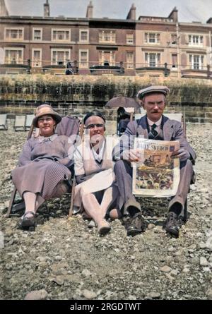 Tre vacanzieri, due donne e un uomo, seduti sulla spiaggia di ciottoli di Hastings, Sussex. Sta leggendo il quotidiano Daily Sketch. Una terrazza di hotel può essere visto sullo sfondo. Foto Stock
