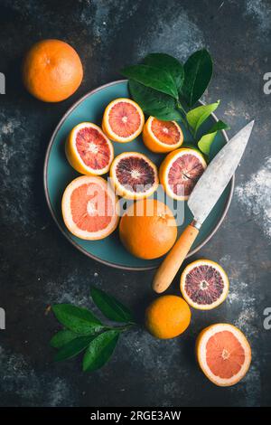 Arance sanguigne e pompelmo rosa, tagliati a metà e interi, con foglie di agrumi, in ciotola di ceramica blu e sul tavolo, con coltello su sfondo scuro Foto Stock