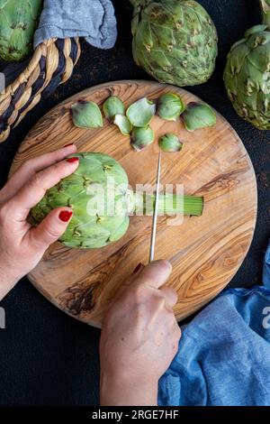 Le mani di una donna tagliano lo stelo di un carciofo su una tavola rotonda di legno. Foto Stock