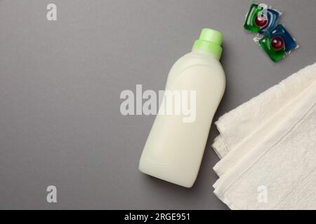 Bottiglia di ammorbidente per tessuti, asciugamani in spugna e cialde di detergente per bucato su sfondo grigio, piatto. Spazio per il testo Foto Stock