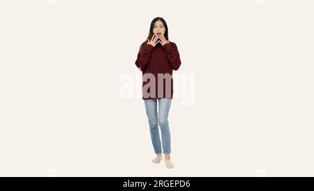 Una bella giovane donna asiatica in un accogliente maglione rosso posa con una faccia scioccata e sbalordita, in piedi su uno sfondo bianco isolato. Foto Stock