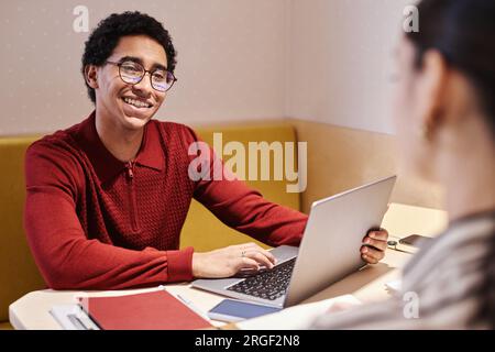 Ritratto di un giovane uomo arabo che indossa occhiali e sorride durante un incontro di lavoro con un collega nello stand dell'ufficio con illuminazione elettrica Foto Stock