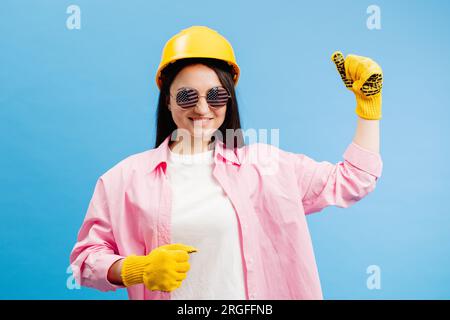 Donna con casco giallo e guanti che puntano e gesturano contro lo sfondo blu dello studio. Foto Stock