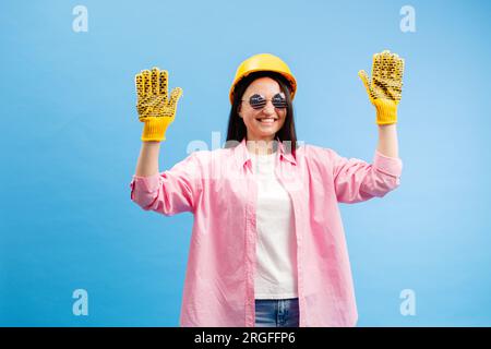 Donna con casco giallo e guanti che puntano e gesturano contro lo sfondo blu dello studio. Foto Stock