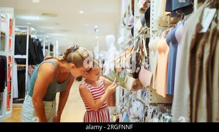 Giovane madre e bambino sceglie i vestiti in boutique di abbigliamento sportivo. Allegro bambino e madre scelta di abbigliamento sportivo nel negozio al dettaglio. Madre e figlia scelgono di cercare vestiti nel centro commerciale Foto Stock