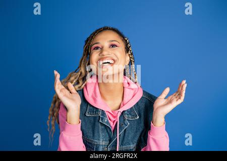 felice donna afro-americana con i dreadlocks che ridono e gestano su sfondo blu, positivo Foto Stock
