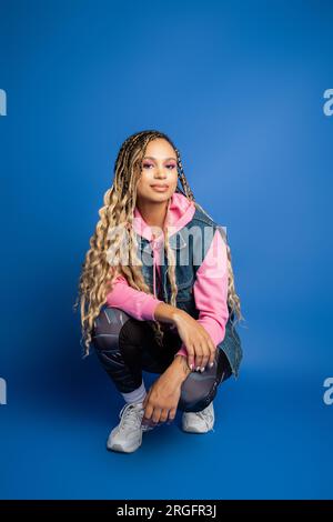 donna dalla pelle scura sicura di sé con dreadlock seduti in un outfit sportivo su sfondo blu, sorridi Foto Stock