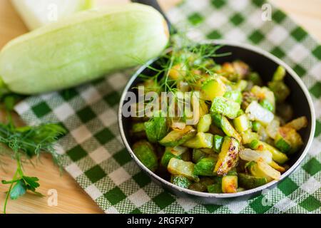 Zucchine a fette fritte con cipolle ed erbe in una padella su un tavolo di legno. Foto Stock