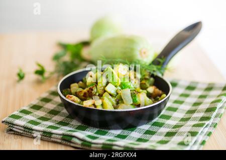 Zucchine a fette fritte con cipolle ed erbe in una padella su un tavolo di legno. Foto Stock