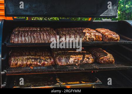 Grande grigliata per fumatori al festival pubblico. Carne e pancetta preparate in un affumicatore per barbecue. Foto Stock