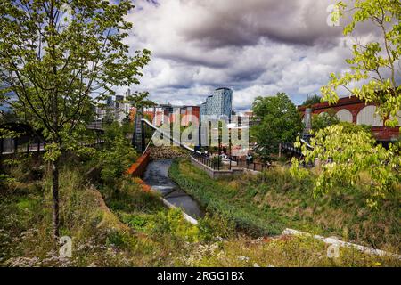 Il fiume Medlock scorre attraverso un parco urbano con area giochi e con lo skyline del centro città sullo sfondo Mayfield Park, Manchester, Inghilterra. Foto Stock