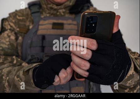 Soldato ucraino in forma di pixel militare e giubbotto antiproiettile con bandiera Ucraina che tiene lo smartphone a mano in guanto nero. Foto Stock