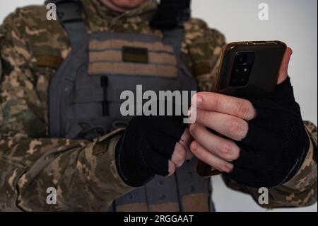 Soldato ucraino in forma di pixel militare e giubbotto antiproiettile con bandiera Ucraina che tiene il telefono a mano in guanto nero. Foto Stock