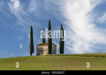 Piccola chiesa circondata da cipressi mediterranei tipici del paesaggio toscano, italiano contro nuvole di piume in un cielo blu con mezza luna appena sopra Foto Stock