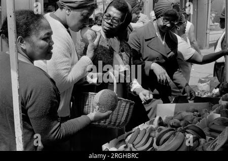 Multietnico Regno Unito anni '1970. Portobello Road, Notting Hill, bancarella del mercato del sabato a Londra. Gruppo di donne nere britanniche che comprano frutta, odorano per vedere se matura. 1975, HOMER SYKES Foto Stock