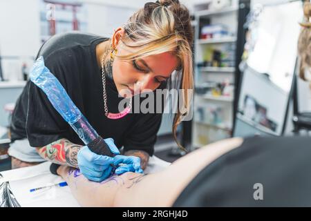 Concentrazione durante il lavoro. Tatuatrice caucasica al lavoro sul suo nuovo disegno del tatuaggio delle gambe. Donna che tiene in mano una pistola per tatuaggio. Foto di alta qualità Foto Stock