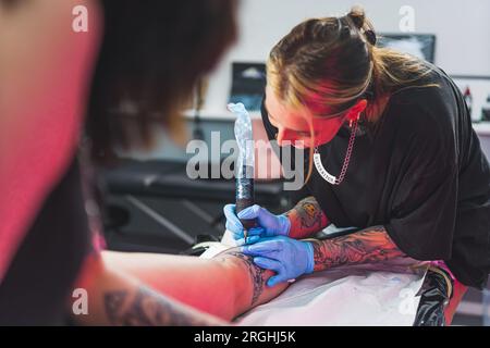 Una tatuatrice donna che disegna un nuovo disegno sul suo cliente femminile. Il lungo processo di tatuaggio delle gambe. Tatuatrice donna felice e positiva. Foto di alta qualità Foto Stock