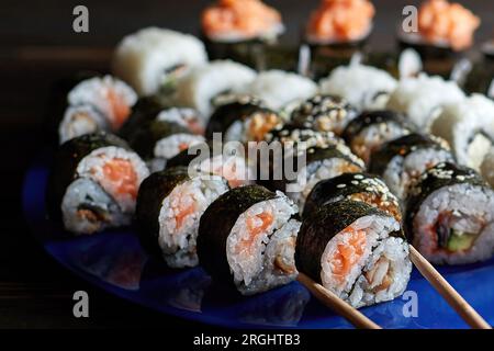 Le belle mani femminili prendono il sushi con le bacchette. Sfondo scuro. Tavola di legno. Vista laterale. Vista orizzontale piastra blu. Sushi. Assortiti Foto Stock